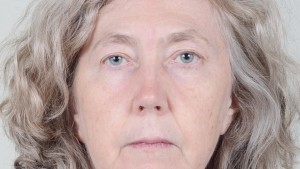 Image of a white woman looking at the camera. She has grey hair and a blue shirt. She is standing infront of a white background